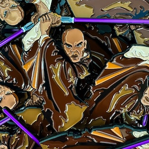 Peut inclure: Épinglette en émail représentant un personnage chauve en robe marron, maniant un sabre laser violet. La broche présente un design détaillé avec des nuances de marron et de beige, avec un contour argenté. Le personnage a une expression sévère.
