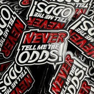 May include: A close-up of several black and white enamel pins with the text "Never Tell Me The Odds!" in red and white lettering. The pins are stacked on top of each other.