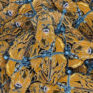 May include: A pile of enamel pins depicting Chewbacca from Star Wars. The pins are predominantly brown and yellow, with blue eyes and silver accents. The pins have a shiny finish and are arranged in a heap.