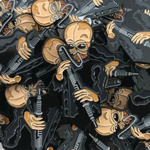 Puede incluir: Primer plano de un montón de pines de esmalte negro con un extraterrestre de dibujos animados sosteniendo un arma. El extraterrestre tiene una piel de color marrón claro y ojos grandes. Los pines están todos en la misma pose y están dispuestos en un patrón aleatorio.