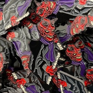 May include: A collection of enamel pins featuring a red demon character in a black suit with a purple tie. The demon has horns and a menacing expression. The pins are all black and red with a glossy finish.