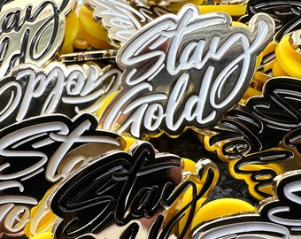 May include: A close-up of a pile of gold and black enamel pins with the words "Stay Gold" written in a stylized font. The pins are arranged in a random pattern and some have a yellow backing.