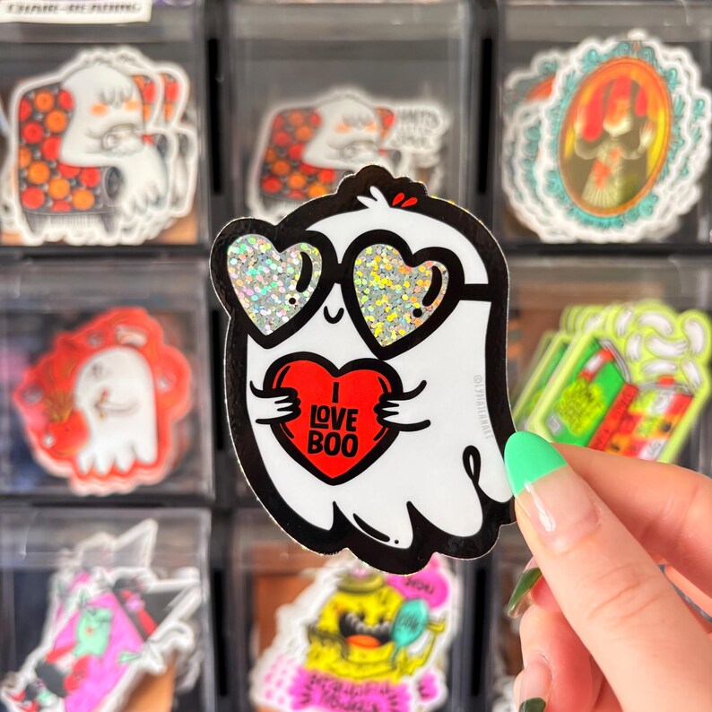 May include: A white ghost sticker with black outlines, wearing heart-shaped glitter sunglasses and holding a red heart with "I LOVE BOO" text. Other ghost-themed stickers are visible in the background.