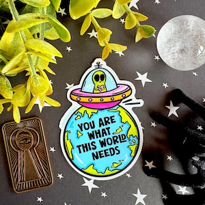 May include: A colorful sticker featuring an alien in a UFO above a globe. The sticker has the text "YOU ARE WHAT THIS WORLD NEEDS". The background is black with white stars and a gold keyhole-shaped charm.