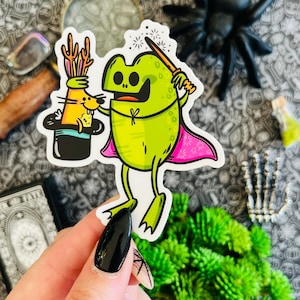 Puede incluir: Una rana verde con una capa rosa y una varita mágica está realizando un truco de magia. Un conejo está en un sombrero de copa. La rana está sonriendo y el conejo parece sorprendido.