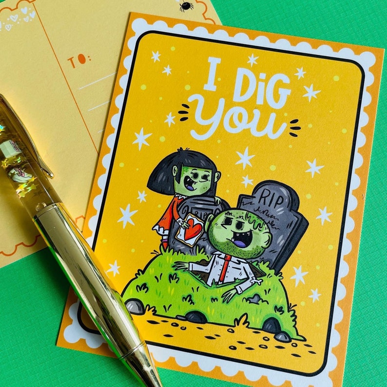 May include: A yellow greeting card with the text "I Dig You" and a cartoon illustration of zombies. The illustration features a zombie emerging from a grave and another holding a heart. A gold pen and a postcard are in the image.