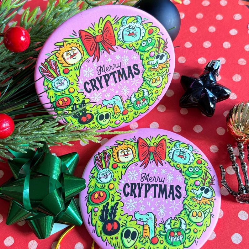 Puede incluir: Dos adornos redondos rosas con un dise&ntilde;o de corona verde y el texto "Merry CRYPTMAS". La corona presenta monstruos de dibujos animados, un lazo rojo y copos de nieve. Los adornos est&aacute;n sobre un fondo de lunares rojos y blancos con decoraciones navide&ntilde;as.