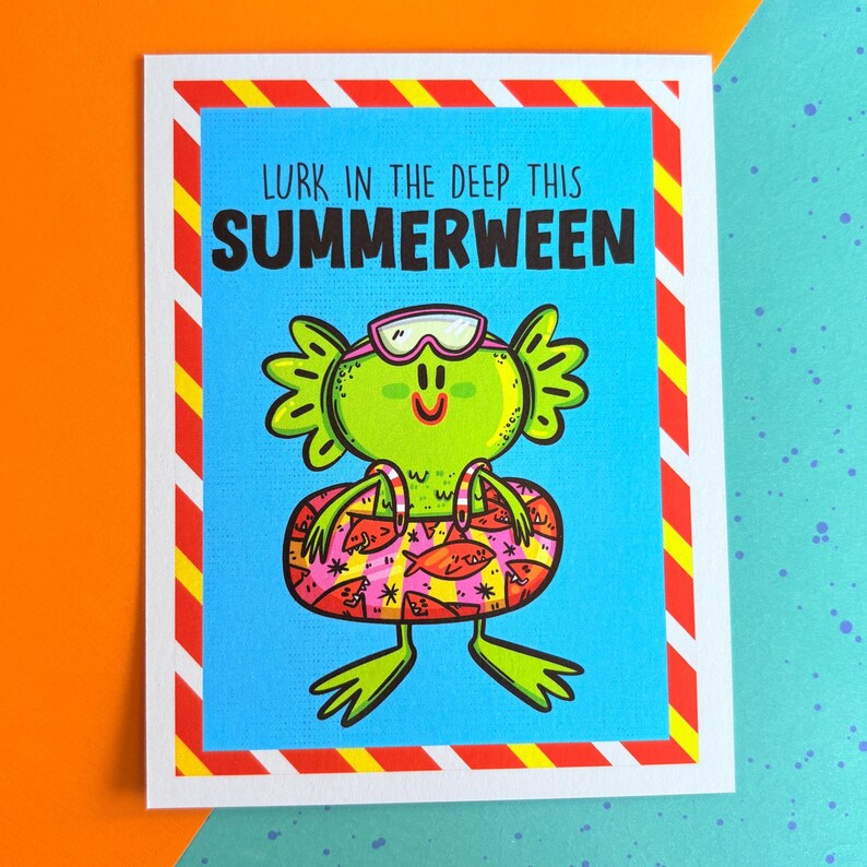 May include: Greeting card with a cartoon illustration of a green creature wearing swim goggles and a pink and yellow inner tube. The card has a blue background with the text "LURK IN THE DEEP THIS SUMMERWEEN" in black.