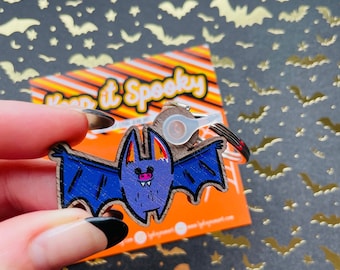 Pode incluir: Um chaveiro de madeira em forma de morcego roxo e azul com um design pintado de branco e laranja. O chaveiro tem um anel prateado e um amuleto de plástico transparente com uma pequena pérola prateada. O texto "Keep it Spooky" é visível em um cartão atrás do chaveiro.