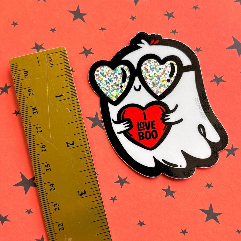 May include: A white ghost sticker with black outlines, wearing heart-shaped glasses with glitter. The ghost holds a red heart with "I LOVE BOO" text. The sticker is on a red background with black stars. A ruler shows inches.