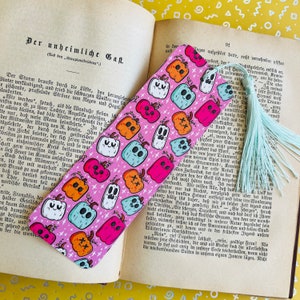May include: A pink bookmark with a repeating pattern of white ghosts and orange, pink, and blue pumpkins. The bookmark has a light blue tassel.