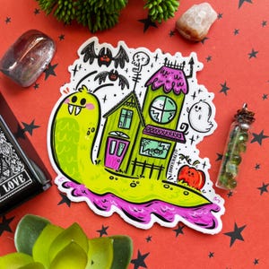 May include: A green cartoon snail carrying a green and pink haunted house with a pink door and a spider web window. The snail has a toothy grin and is surrounded by bats and stars.