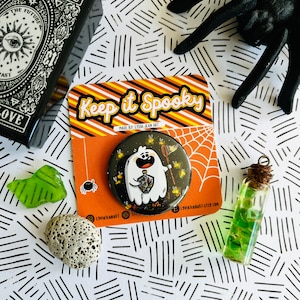 May include: A black and white button with a cartoon ghost holding a jar of fireflies. The button is on a card with the text "Keep it Spooky" and "Made by Lydia Jean Art".
