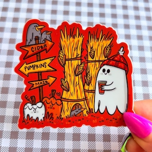 May include: A fall-themed sticker featuring a cartoon ghost wearing a red beanie and holding a coffee cup. The sticker includes a signpost with arrows pointing to cider, pumpkins, and a maze, along with pumpkins, hay bales, and corn.