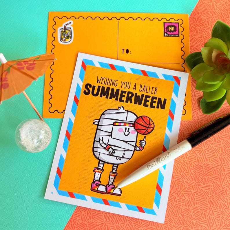 May include: A greeting card with the text "Wishing you a baller Summerween" features a cartoon mummy holding a basketball. The card has a yellow background with blue and red stripes. An orange envelope and a pen are also in the image.
