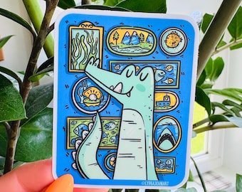 Könnte beinhalten: Ein blauer Aufkleber mit einem Cartoon-Alligator mit hellgrünem Körper und lächelndem Gesicht. Der Aufkleber hat verschiedene gerahmte Bilder von Unterwasserszenen, darunter Fische, ein U-Boot und ein Hai. Der Text "OLYDIAJEANART" befindet sich unten rechts.