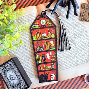 Puede incluir: Un marcapáginas negro con un diseño de estantería roja, con libros, pociones y una bola de cristal. También se representa un gato negro, una telaraña y una calavera. Una borla negra cuelga de la parte superior. El marcapáginas está en un libro abierto.