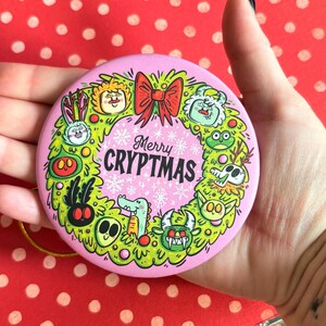 Puede incluir: Un adorno navide&ntilde;o redondo y rosa con un dise&ntilde;o de corona verde. La corona presenta monstruos de dibujos animados y un lazo rojo. El texto "Merry CRYPTMAS" est&aacute; en el centro, rodeado de copos de nieve. Se adjunta una cuerda dorada para colgar.
