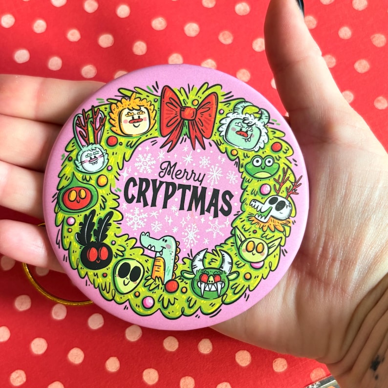 Puede incluir: Un adorno navide&ntilde;o redondo y rosa con un dise&ntilde;o de corona verde. La corona presenta monstruos de dibujos animados y un lazo rojo. El texto "Merry CRYPTMAS" est&aacute; en el centro, rodeado de copos de nieve. Se adjunta una cuerda dorada para colgar.