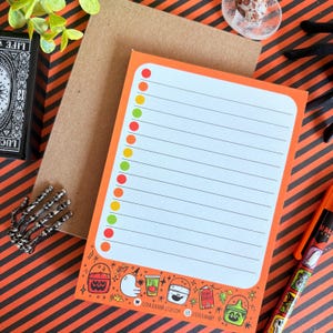 May include: Orange Halloween-themed notepad with a white interior and colorful dots along the left side. The bottom border features Halloween illustrations. A brown envelope is behind the notepad. A pen with Halloween graphics is visible.