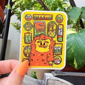 May include: A yellow sticker featuring a cartoon Bigfoot with orange fur and a smiling face. The sticker has various framed illustrations of trees, mountains, and a UFO. The word "CHUNKY" is visible on the sticker.