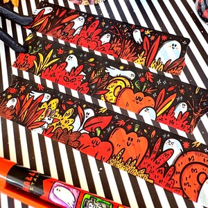 Pu&ograve; includere: Tre segnalibri illustrati a tema Halloween. Ogni segnalibro presenta uno sfondo nero con strisce bianche e illustrazioni di fantasmi, zucche e fogliame autunnale in arancione, rosso e giallo. Un nastro nero &egrave; attaccato.