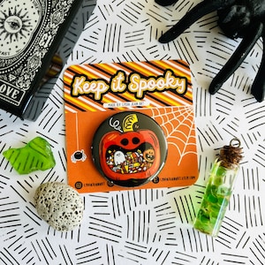 May include: A round button with a black background and a cartoon pumpkin with a smiling face. The pumpkin is filled with candy, ghosts, and other Halloween treats. The button says "Keep it Spooky" and "Made by Lydia Jean Art".