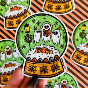 May include: A green and yellow snow globe sticker with three ghosts singing Christmas carols inside. The ghosts are white with black eyes and mouths. The snow globe is surrounded by a white border and has a black spiderweb design on the base.
