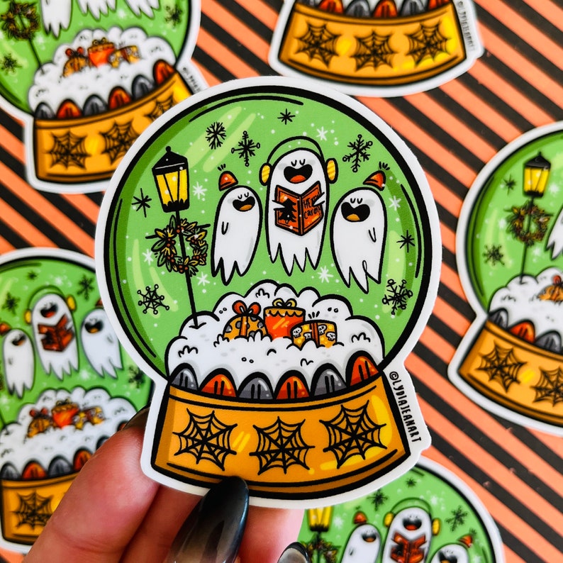 May include: A green and yellow snow globe sticker with three ghosts singing Christmas carols inside. The ghosts are white with black eyes and mouths. The snow globe is surrounded by a white border and has a black spiderweb design on the base.