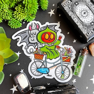 May include: A sticker featuring a green and red character riding a bicycle with a unicorn and flowers in baskets. The character is wearing a green cloak and has a red face with two large eyes. The sticker is on a white background.