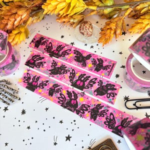 Puede incluir: Tres tiras de cinta washi rosa con un patr&oacute;n repetitivo de criaturas negras similares a polillas con ojos de color rosa brillante, rodeadas de peque&ntilde;as estrellas y garabatos amarillos y rosas. El texto LYDIAJEANART est&aacute; impreso.