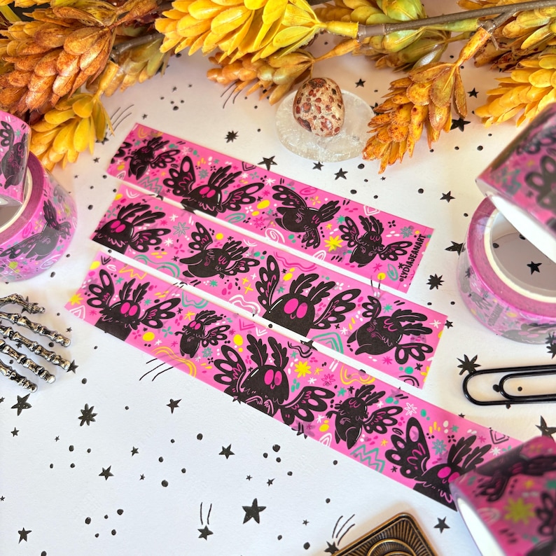 Puede incluir: Tres tiras de cinta washi rosa con un patr&oacute;n repetitivo de criaturas negras similares a polillas con ojos de color rosa brillante, rodeadas de peque&ntilde;as estrellas y garabatos amarillos y rosas. El texto LYDIAJEANART est&aacute; impreso.