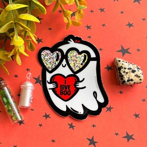 May include: A white ghost sticker with heart-shaped sunglasses and a red heart that says "I Love Boo." The sticker is on a red background with black star shapes. A small decorative bottle and a stone are also visible.