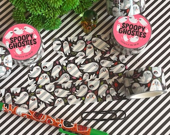 Puede incluir: Rollos de cinta washi con fondo negro, ilustraciones de fantasmas blancos y el texto "SPOOPY GHOSTIES" en las etiquetas. También se incluyen un bolígrafo naranja con pequeños iconos gráficos y un clip de papel negro.