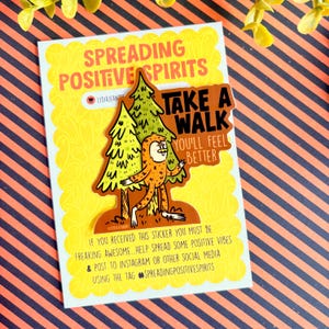 Puede incluir: Una pegatina con el texto "SPREADING POSITIVE SPIRITS" y un gr&aacute;fico de Bigfoot. El Bigfoot es naranja y marr&oacute;n, caminando cerca de dos &aacute;rboles verdes. La pegatina tambi&eacute;n dice "TAKE A WALK YOU'LL FEEL BETTER".