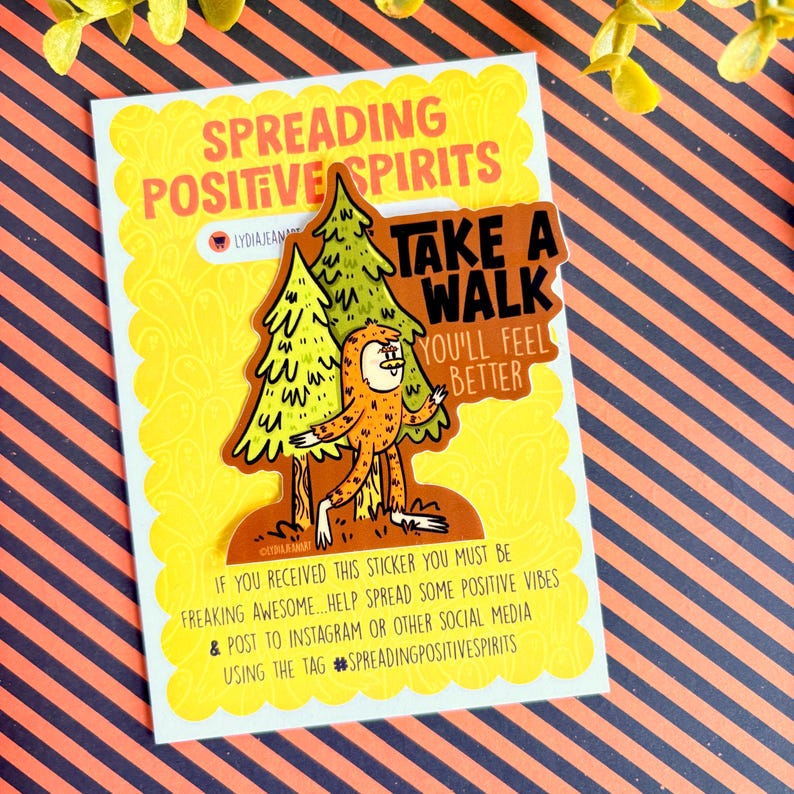 Puede incluir: Una pegatina con el texto "SPREADING POSITIVE SPIRITS" y un gr&aacute;fico de Bigfoot. El Bigfoot es naranja y marr&oacute;n, caminando cerca de dos &aacute;rboles verdes. La pegatina tambi&eacute;n dice "TAKE A WALK YOU'LL FEEL BETTER".