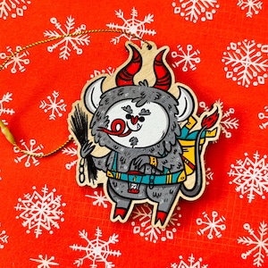 May include: A wooden Christmas ornament shaped like a gray monster with red horns, a red nose, and a blue and yellow backpack. The monster is holding a black stick in its right hand.