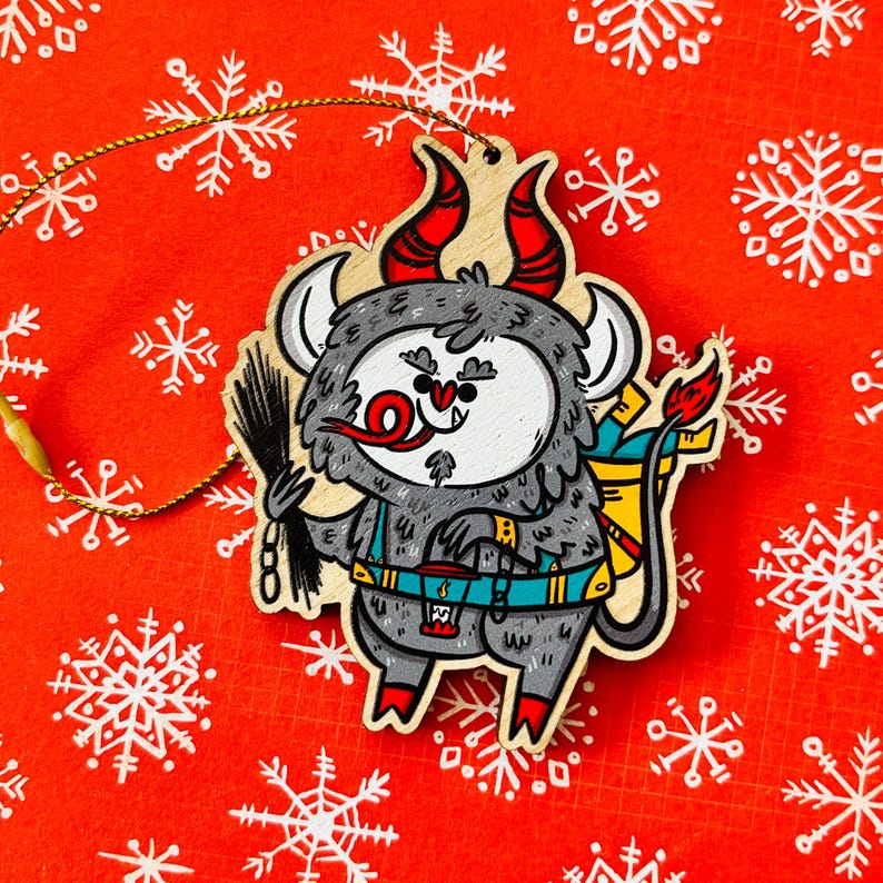 May include: A wooden Christmas ornament shaped like a gray monster with red horns, a red nose, and a blue and yellow backpack. The monster is holding a black stick in its right hand.