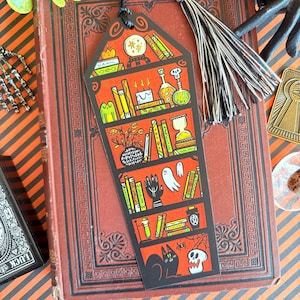 以下が含まれることがあります： 黒いタッセルが付いたハロウィーンをテーマにしたブックマーク。さまざまな不気味な要素を備えた赤い本棚のデザインが特徴です。棚には本、ポーション、クリスタルボール、黒猫、スカル、幽霊が表示されています。ブックマークはヴィンテージの本の上にあります。
