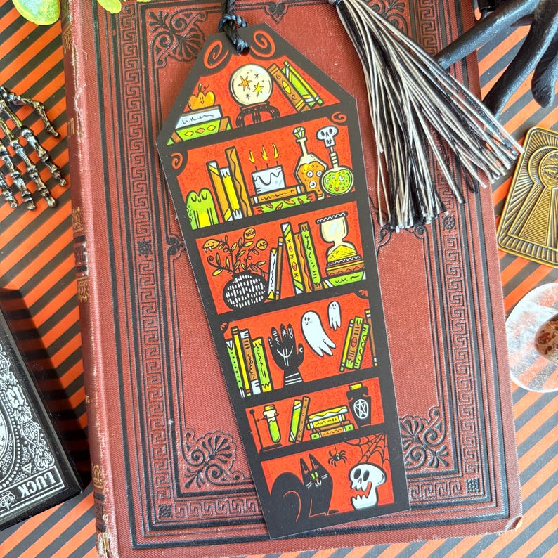 以下が含まれることがあります： 黒いタッセルが付いたハロウィーンをテーマにしたブックマーク。さまざまな不気味な要素を備えた赤い本棚のデザインが特徴です。棚には本、ポーション、クリスタルボール、黒猫、スカル、幽霊が表示されています。ブックマークはヴィンテージの本の上にあります。