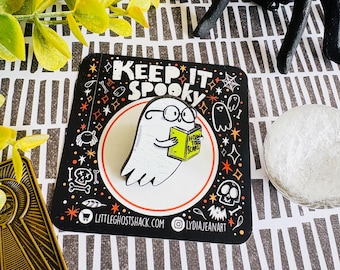 Puede incluir: Un pin negro y blanco con el texto "KEEP IT SPOOKY" y un fantasma de dibujos animados leyendo un libro titulado "HOW TO SCARE". El pin tiene un diseño de fantasma blanco con gafas, rodeado de estrellas naranjas y blancas y otros elementos espeluznantes.