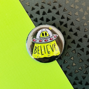 May include: A green alien in a flying saucer with the word "BELIEVE" written on the side. The saucer is yellow with red accents and white stars.