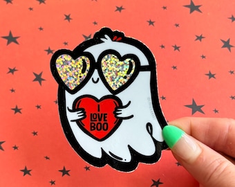 May include: A white ghost sticker with black outlines, wearing heart-shaped sunglasses with glitter. The ghost is holding a red heart with the words "I LOVE BOO" in white. The background is orange with black star shapes.