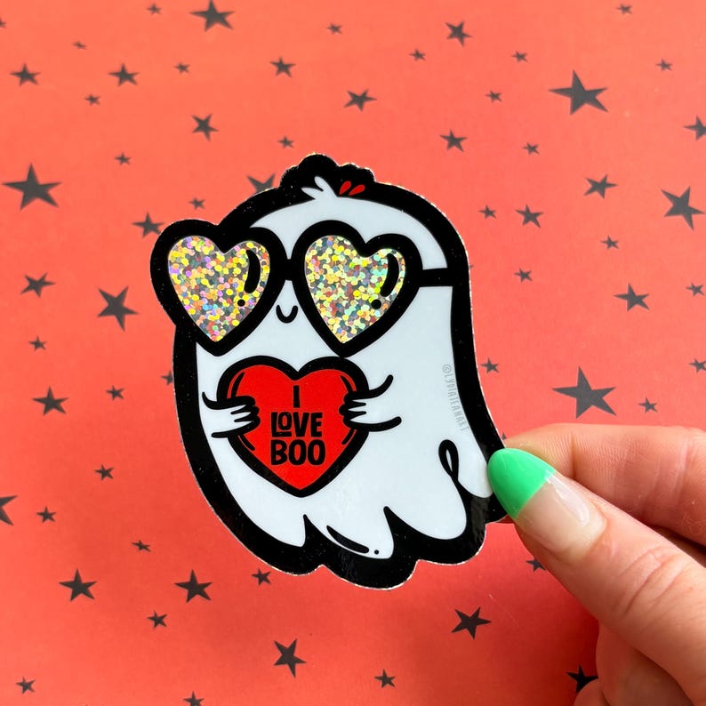 May include: A white ghost sticker with black outlines, wearing heart-shaped sunglasses with glitter. The ghost is holding a red heart with the words "I LOVE BOO" in white. The background is orange with black star shapes.