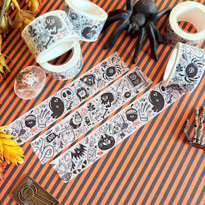 May include: Halloween-themed decorative tape rolls with black and white illustrations of ghosts, skulls, and spiders. The tape features the text "Creep Real" and other spooky graphics. The background is orange and black striped.