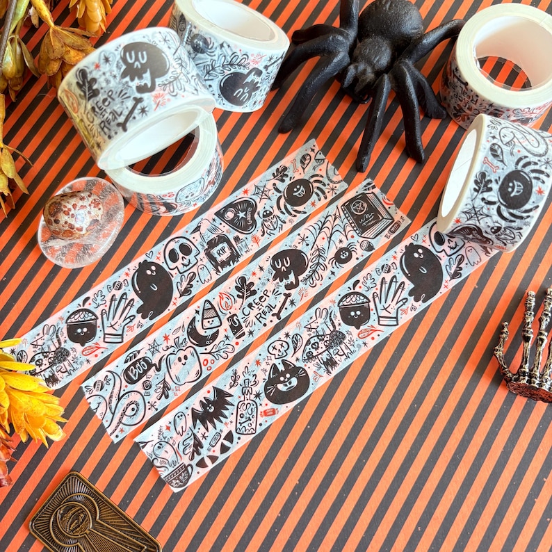 May include: Halloween-themed decorative tape rolls with black and white illustrations of ghosts, skulls, and spiders. The tape features the text "Creep Real" and other spooky graphics. The background is orange and black striped.