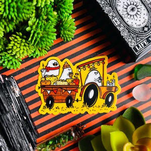 Puede incluir: Una pegatina amarilla con una ilustración de dibujos animados de tres fantasmas montando en un carro decorado. El carro está decorado con luces y tiene un letrero que dice "¡Boo!"