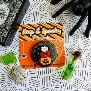 May include: A round, black Halloween button with three stacked jack-o'-lanterns, the top one white. The button is on a card that says "Keep it Spooky." A small spider and a spiderweb are also visible.