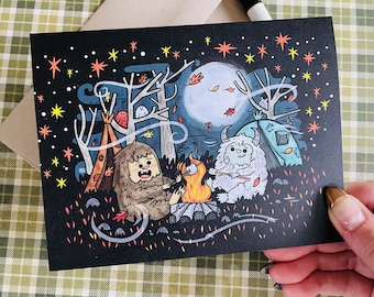 Könnte beinhalten: Eine schwarze Karte mit einer weißen und blauen Illustration von zwei pelzigen Kreaturen, die um ein Lagerfeuer unter einem sternenklaren Nachthimmel sitzen. Die Kreaturen sind ein Yeti und ein Sasquatch.