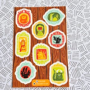 Könnte beinhalten: Eine Holztafel zeigt neun gerahmte Illustrationen verschiedener Kreaturen. Jeder Rahmen hat eine andere Farbe, und die Illustrationen zeigen einen Löwen, ein Kaninchen, einen Drachen und eine Fledermaus. Der Social-Media-Handle und die Website des Künstlers sind unten.